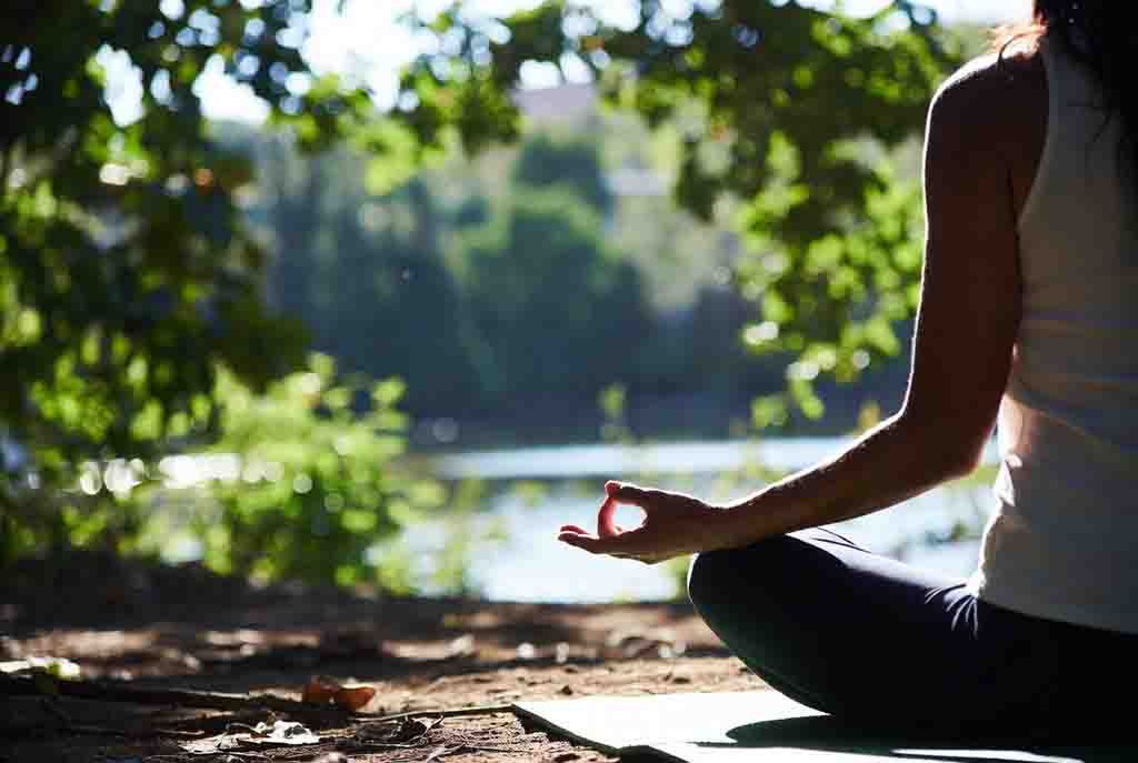 Frau meditiert in der Natur am Wasser in der Morgensonne während eines erholsamen Wellnessurlaubs