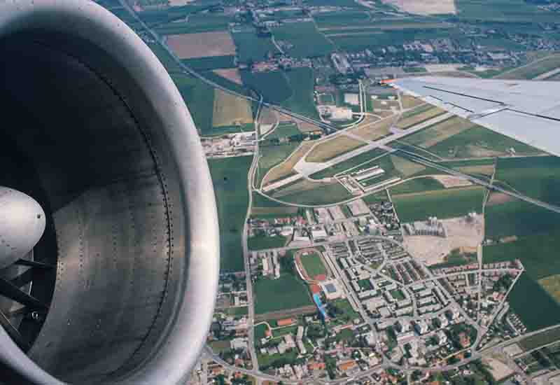 Nahaufnahme eines großen Flugzeugtriebwerks und eines Teils der Tragfläche im Vordergrund, mit Blick aus dem Fenster auf eine weitläufige Landschaft, Wohngebiete, Felder und ein Stadion tief unter dem Flugzeug.