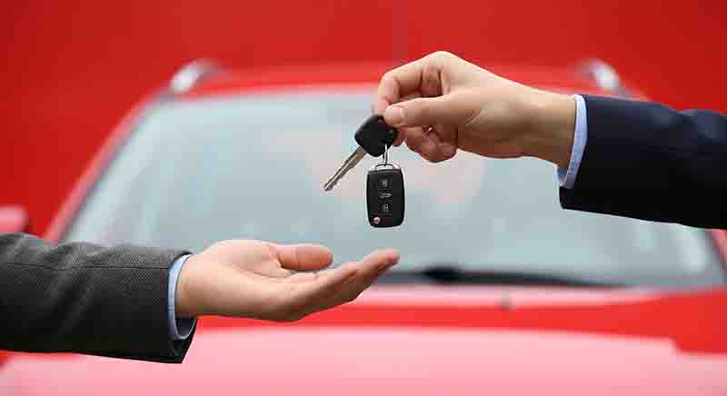 Nahaufnahme von zwei Händen, die Autoschlüssel vor einem roten Auto austauschen. Der Hintergrund ist unscharf, wobei das leuchtende Rot des Autos dominiert. Das Bild deutet auf das Mieten oder Abholen eines Autos hin.